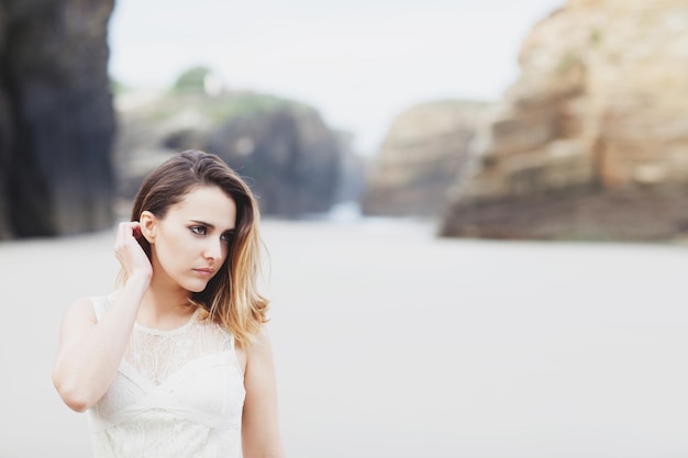 Portret van een mooie vrouw in een romantische jurk raakt haar haar aan op een strand met hoge kliffen