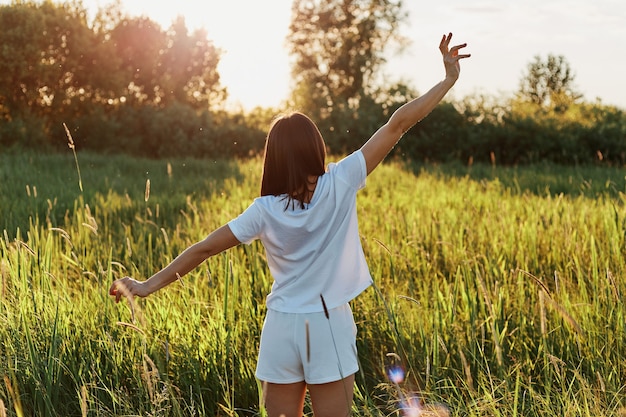 Portret van een mooie vrouw in witte kleren verheugt zich in de groene weide, achterover staand met wijd gespreide armen, genietend van de schoonheid en rust van de natuur en het zonsonderganglicht.