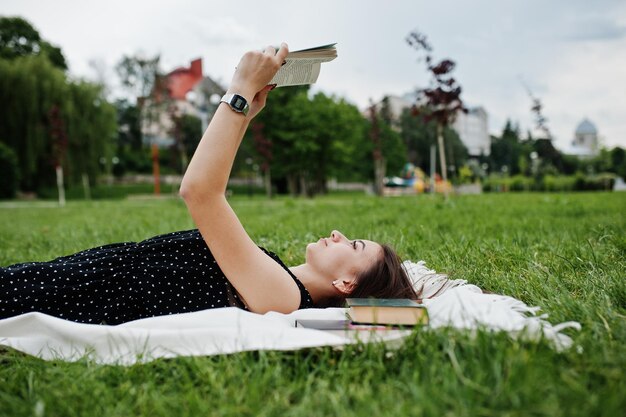 Foto portret van een mooie vrouw in zwarte polka dot-jurk die op de deken op het gras ligt en leest