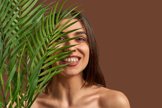 Foto portret van een mooie vrouw op een bruine achtergrond die een tak van een palmboom vasthoudt en glimlacht schoonheidsconcept