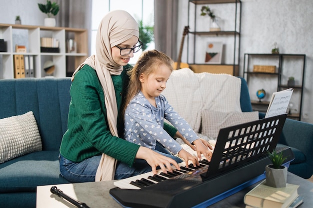 Portret van een moslimleraar in hijab en haar kleine leerling die thuis piano speelt