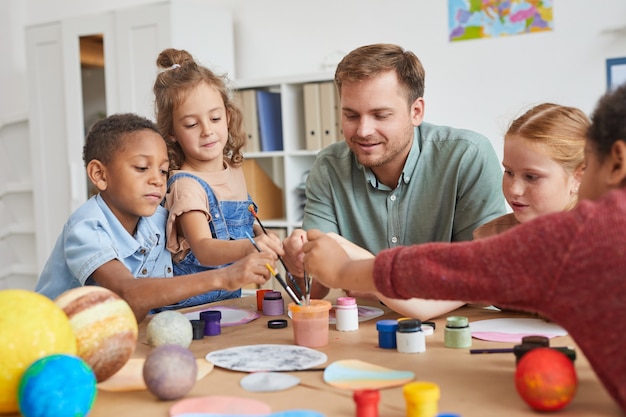 Portret van een multi-etnische groep kinderen die borstels vasthouden en planeetmodel schilderen terwijl ze genieten van kunst- en ambachtlessen op school of ontwikkelingscentrum