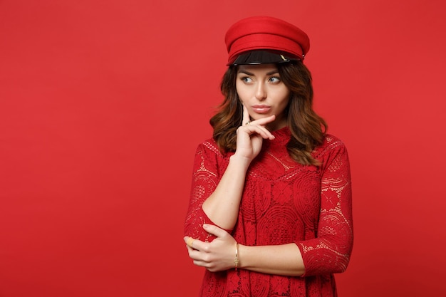 Portret van een nadenkende jonge vrouw in kanten jurk, pet opzij kijkend, handsteun op de kin geïsoleerd op een heldere rode achtergrond in de studio. Mensen oprechte emoties, lifestyle concept. Bespotten kopie ruimte.