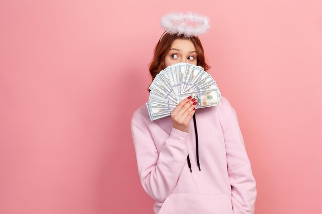 Portret van een nieuwsgierig tienermeisje met krullend haar in hoodie met halo op hoofd dat gezicht verbergt met een stel dollarbankbiljetten en naar de kant kijkt. Indoor studio-opname geïsoleerd op roze background