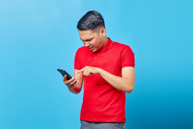 Portret van een ongelukkige knappe Aziatische jongeman die smartphone gebruikt en sms't op blauwe achtergrond
