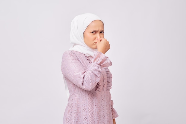 Portret van een ongelukkige, mooie jonge Aziatische moslimvrouw die een hijab en een paarse jurk draagt en walgt terwijl ze haar neus vasthoudt om te voorkomen dat ze vies ruikt geïsoleerd op een witte studio achtergrond