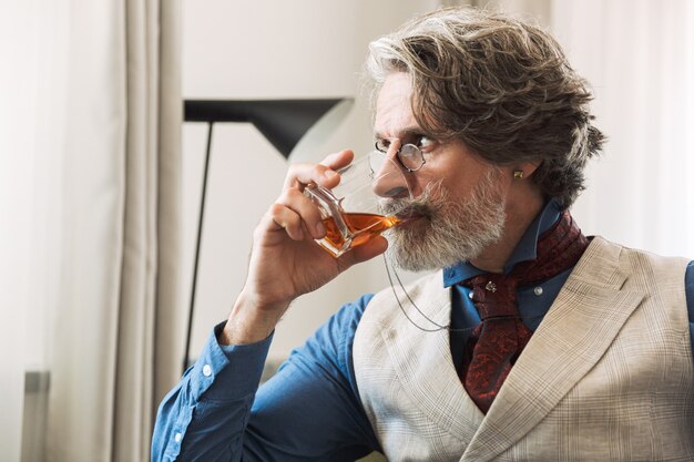 Portret van een ongeschoren volwassen professor met een stijlvol pak en een bril die alcohol uit glas drinkt terwijl hij op een fauteuil in een hotelappartement zit