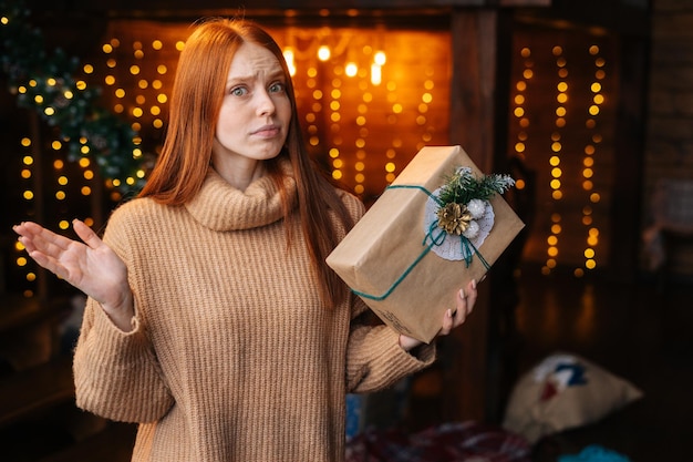 Portret van een onrustige roodharige jonge vrouw die een mooie geschenkdoos met kerstcadeau in de hand houdt