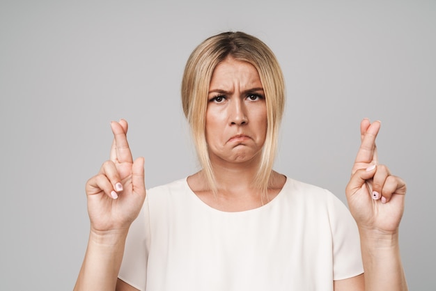 Portret van een ontevreden trieste mooie jonge blonde vrouw poseren geïsoleerd over grijze muur gekleed in basic wit t-shirt, maakt hoopvol gebaar met gekruiste vingers