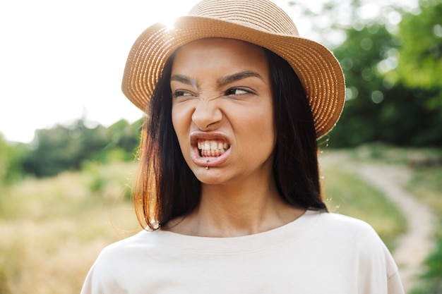 Portret van een ontevreden vrouw met een strohoed en lippiercing die haar tanden laat zien terwijl ze in een groen park wandelt