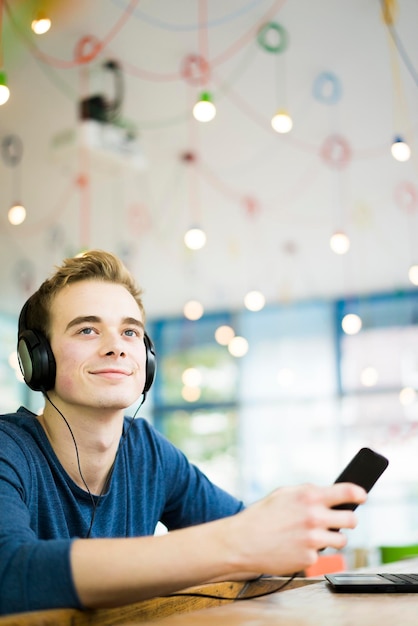 Portret van een ontspannen jonge man die muziek luistert met een koptelefoon en een smartphone in een coffeeshop