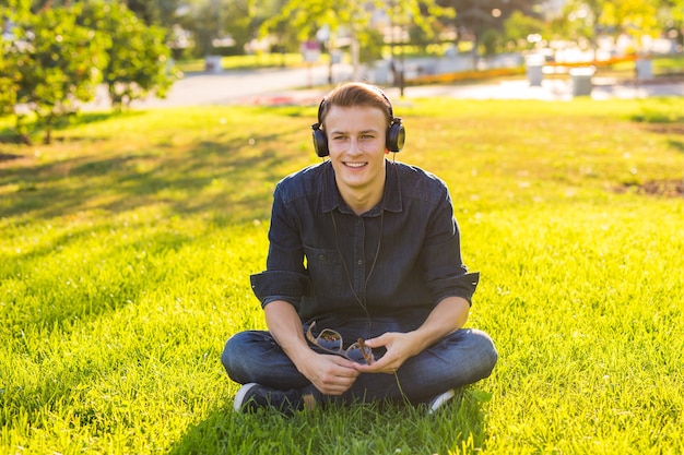 Portret van een ontspannen jonge man zittend op het gras in het park en luisteren naar muziek op een koptelefoon.