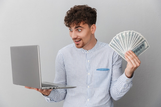 Portret van een opgetogen man gekleed in hemd terwijl hij een zilveren laptop vasthoudt en een hoop geld cash geïsoleerd over grijze muur