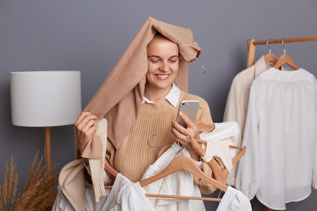 Portret van een opgetogen tevreden vrouw shopper met kleding op haar hoofd met kleding op hangers die met smartphone in handen staan en naar het display kijken met een brede glimlach