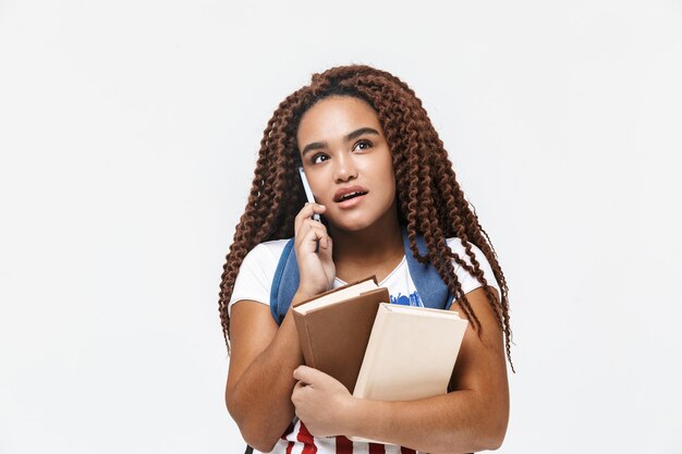 Portret van een opgetogen vrouw met een rugzak die op een mobiele telefoon praat terwijl ze studerende boeken vasthoudt die tegen een witte muur zijn geïsoleerd