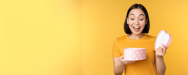 Portret van een opgewonden aziatische vrouw met een open geschenkdoos met een verrast blij gezicht over een gele achtergrond