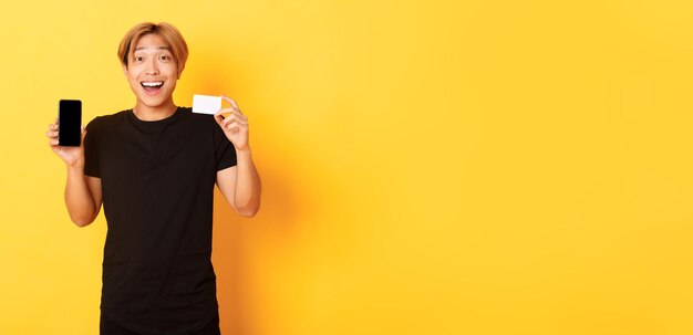 Portret van een opgewonden, gelukkige aziatische man die het scherm van de mobiele telefoon en een creditcard met een vrolijke glimlach toont