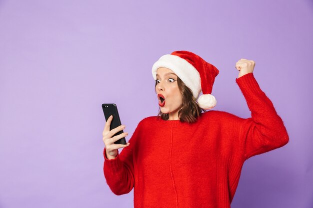 Portret van een opgewonden gelukkige jonge vrouw met kerstmuts geïsoleerd over paarse muur met behulp van mobiele telefoon.