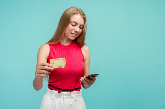 Portret van een opgewonden jong meisje dat plastic creditcard toont terwijl hij mobiele telefoon vasthoudt die over blauwe achtergrond wordt geïsoleerd