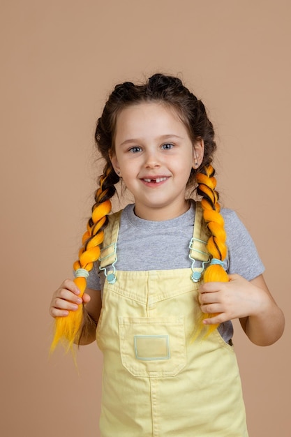 Portret van een opgewonden jong meisje met gele kanekalon-vlechten met handen glimlachend met ontbrekende tand kijkend naar camera met gele jumpsuit en grijze tshirt op beige achtergrond