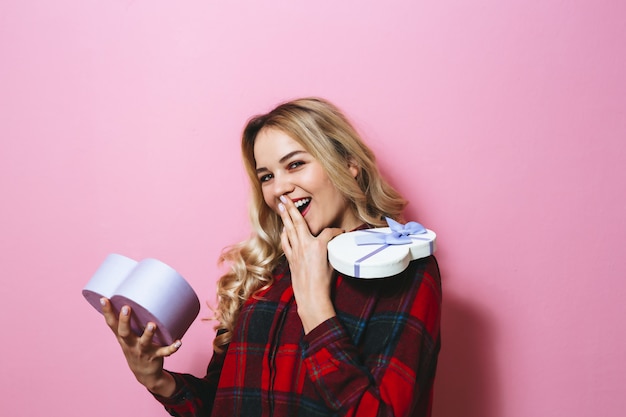 Portret van een opgewonden jong mooi blondemeisje die een gift houden en gelukkig over roze achtergrond