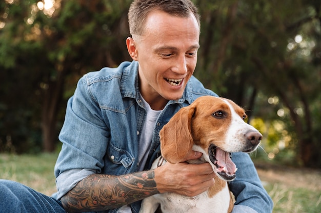 Portret van een opgewonden jongeman in spijkerkleding die lacht en zijn hond knuffelt in het park