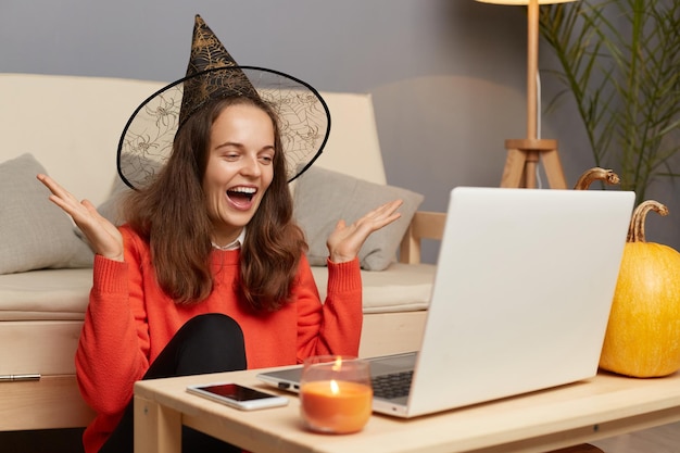 Portret van een opgewonden positieve vrouw met een heksenhoed die vooraan op een laptop in het interieur zit en een videogesprek heeft of een livestream uitzendt met opgeheven armen in opwinding