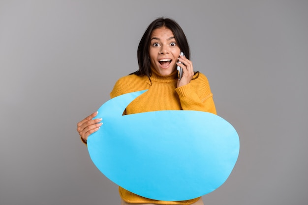 Portret van een opgewonden toevallige jonge vrouw die over grijze muur wordt geïsoleerd, die op mobiele telefoon spreekt, lege toespraakbel toont