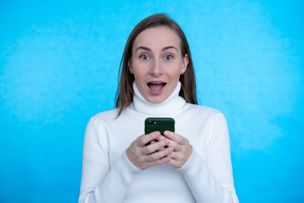 Portret van een opgewonden vrouw die en mobiele telefoon bevindt die over blauwe muur wordt geïsoleerd.