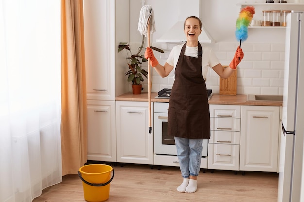 Portret van een opgewonden vrouw met een bruine schort die snel huishoudelijk werk maakt en de vloer afveegt en wast in de moderne keuken die in een goed humeur is met opgeheven armen met haar apparatuur die haar schoonmaak afmaakt