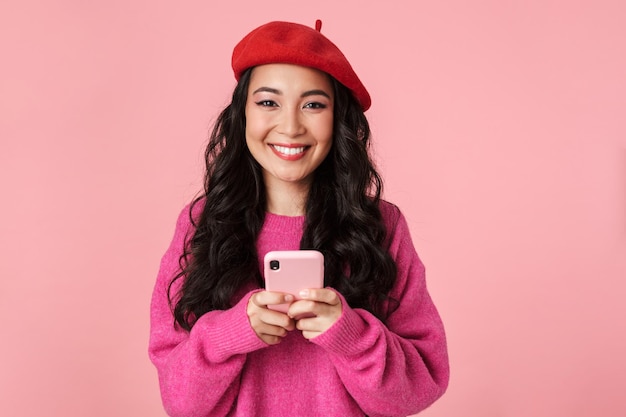 Portret van een optimistisch, mooi Aziatisch meisje met lang donker haar met een baret die lacht en een smartphone vasthoudt die op roze is geïsoleerd