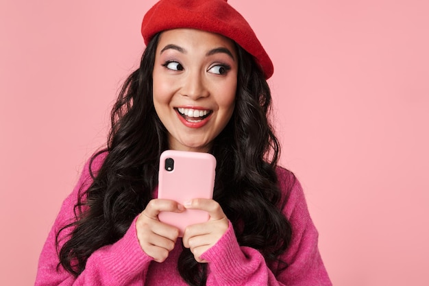 Portret van een optimistisch, mooi Aziatisch meisje met lang donker haar met een baret die lacht en een smartphone vasthoudt die op roze is geïsoleerd