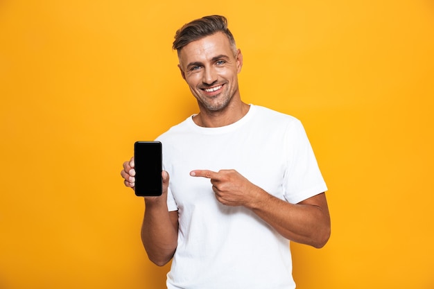 Portret van een optimistische man van 30 in een wit t-shirt die lacht en een mobiele telefoon vasthoudt terwijl hij geïsoleerd op geel staat