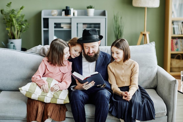 Portret van een orthodox joods gezin met vader die een religieus boek leest aan drie kinderen in huis