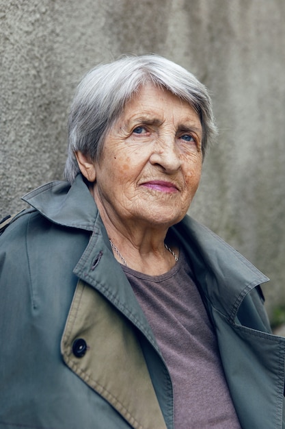 Foto portret van een oude oma van 90 met grijs haar staat tegen grijze muur op straat