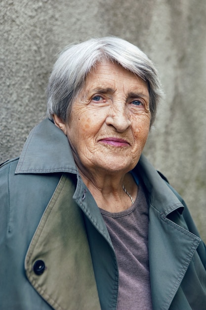 Foto portret van een oude oma van 90 met grijs haar staat tegen grijze muur op straat