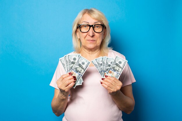 Portret van een oude vriendelijke vrouw in casual t-shirt en een bril met geld in haar handen op een geïsoleerde blauwe muur. emotioneel gezicht. concept rijkdom, win, krediet, pensioen
