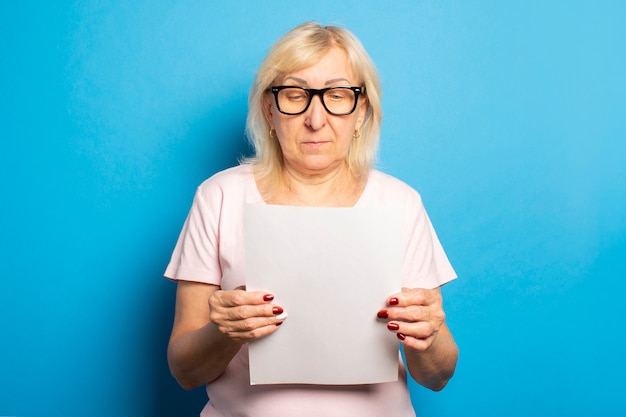 Portret van een oude vriendelijke vrouw in een casual T-shirt en een bril met een vel papier in haar handen en kijken naar het op een geïsoleerde blauwe muur. Emotioneel gezicht. Concept brief, aankondiging