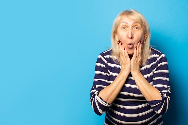Portret van een oude vriendelijke vrouw met een verbaasd gezicht in een casual t-shirt houdt haar handen op haar kin op een geïsoleerde blauwe muur. Emotioneel gezicht. Gebaar van verbazing, schok