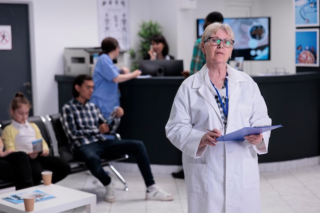 Portret van een oudere arts met een stethoscoop die klembord vasthoudt met patiëntgegevens bij de ziekenhuisreceptie die naar de camera kijkt. Zelfverzekerde senior medic in de lobby van de kliniek, wachtend op de afspraak van de patiënt.