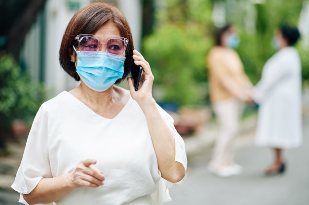 Portret van een oudere Aziatische vrouw met een medisch masker die telefonisch met een familielid praat tijdens het wandelen in het park van het verpleeghuis