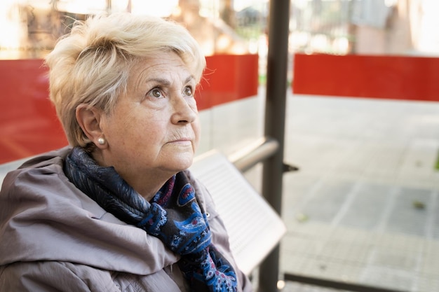 Portret van een oudere blonde vrouw die op de bushalte zit Ernstige oudere dame die wacht op het openbaar vervoer