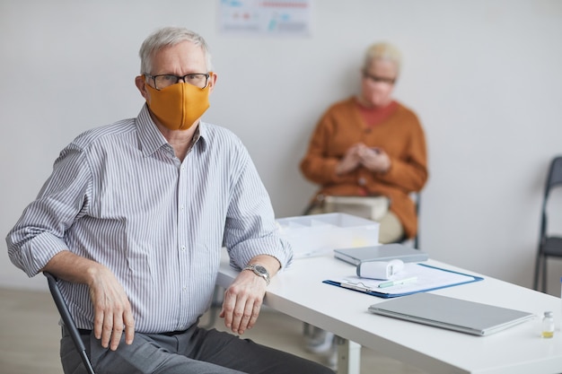 Portret van een oudere man die een masker draagt en naar de camera kijkt terwijl hij aan het bureau van de dokter zit, kopieer ruimte