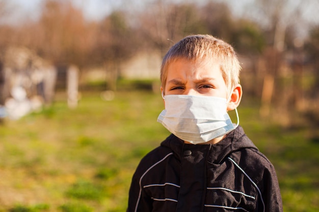 Portret van een overstuur blonde voorschoolse jongen met een beschermend medisch masker buiten in de tuin in de buurt van zijn huis Thuisisolatie en wandelingen in een beschermingsmasker Bescherming tegen virus- en infectiebeheersingsconcept
