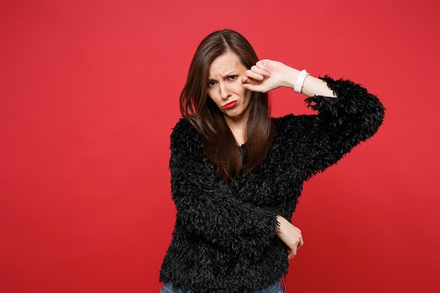 Portret van een overstuur huilende jonge vrouw in een zwarte vachttrui die staat en tranen afveegt geïsoleerd op een felrode muurachtergrond in de studio. Mensen oprechte emoties, lifestyle concept. Bespotten kopie ruimte.