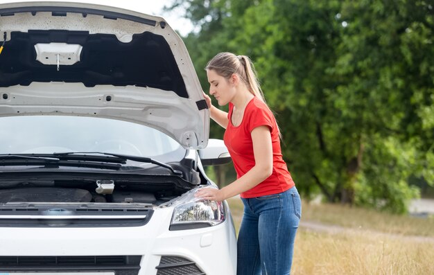 Portret van een overstuur vrouw die onder de motorkap van een oververhitte auto in het veld kijkt