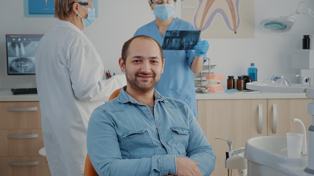 Portret van een patiënt die in de tandartsstoel zit in de mondzorgkliniek, naar de camera kijkt en glimlacht. Blanke man met stomatologische afspraak met tandheelkunde specialist in kabinet.