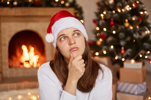 Portret van een peinzende droevige vrouw met een witte trui en een kerstmuts in een ingerichte woonkamer, wegkijkend, kin vasthoudend, denkend, kersttijd.