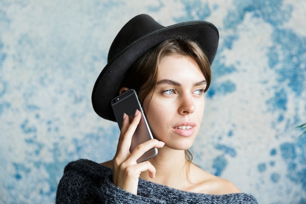 Portret van een peinzende jonge vrouw gekleed in hoed en trui close-up praten op mobiele telefoon over blauwe muur