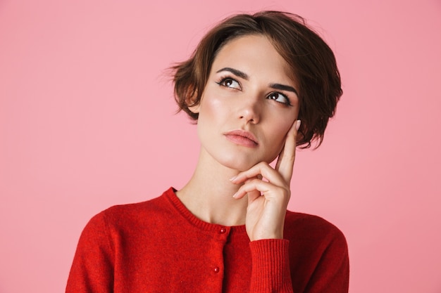 Portret van een peinzende mooie jonge vrouw die rode kleren draagt die zich geïsoleerd over roze achtergrond bevinden